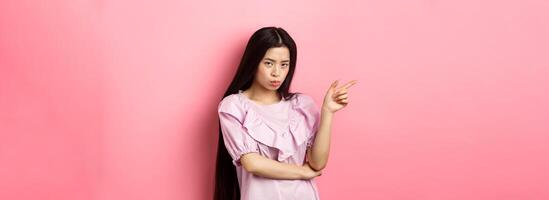 Sad and grumpy asian girl sulking, pointing left and look at camera jealous, want something, complaining and showing desired thing, standing on pink background photo