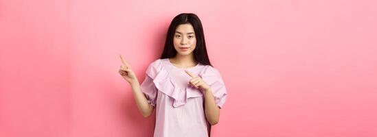 Cheeky asian girl showing interesting advertisement, pointing fingers right at logo and smiling cunning, hinting on good deal, pink background photo