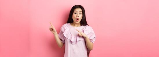 Excited asian woman look amazed, pointing right and gasping fascinated, checking out advertisement, standing on pink background photo