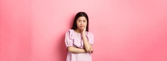 Bored asian teen girl look indifferent at camera, lean face on hand in skeptical pose, standing reluctant against pink background photo