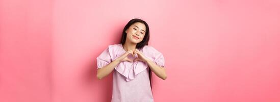 Valentines day concept. Beautiful korean teenager in dress showing heart gesture and smiling with admiration and affection, falling in love, feel romantic, pink background photo