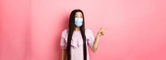 Coronavirus, quarantine and lifestyle concept. Excited asian teen girl in face mask pointing, looking left at logo with amazed face, checking out promotion, white background photo
