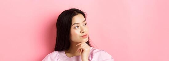 Close-up portrait of romantic asian girl looking aside at logo, thinking or daydreaming, standing against pink background photo