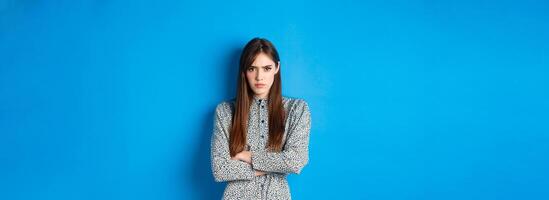 Angry frowning woman in dress, cross arms on chest and feel offended about something unfair, standing upset against blue background photo