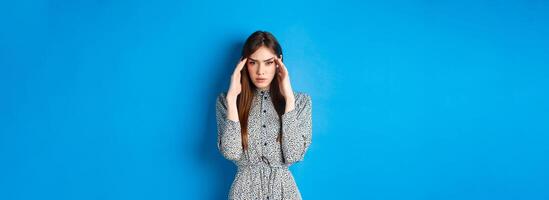 Woman trying to concentrate, touching head and squinting at camera, thinking or remember something, standing in dress on blue background photo