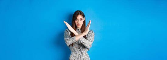 Serious caucasian woman in dress tell no, show cross stop gesture and looking confident, making warning sign, prohibit something bad, standing on blue background photo