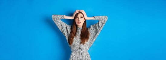 Troubled young woman with long hair, holding hands on head in panic, looking up nervous, having problem, standing on blue background photo