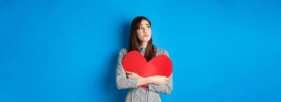 Valentines day. Sad heartbroken girl sobbing, looking aside at empty space and crying upset, hugging big red heart, feeling lonely without lover, standing alone on blue background photo
