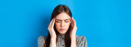 Close-up portrait of young woman feel sick, touching head temples and frowning from headache, having migraine, standing on blue background photo