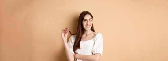 Romantic pensive girl thinking, looking left at empty space and playing with hair strand, smiling dreamy, standing in white dress on beige background photo