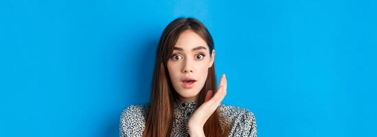Candid girl with long hair and makeup, looking shocked and surprised, open mouth and stare speechless at camera, standing on blue background photo