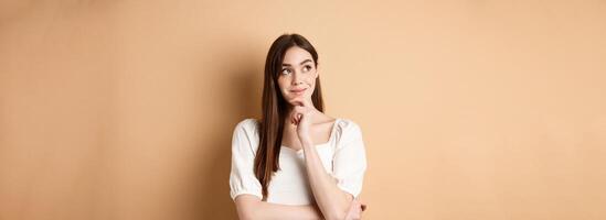 Very interesting. Pensive cute girl thinking and looking at upper left corner logo, smiling pleased, having good idea, standing on beige background photo