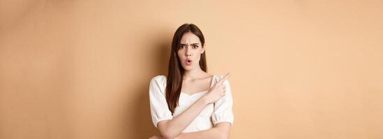 What is that. Shocked and angry woman scolding person, frowning and pointing left with confused face, waiting for explanation, standing on beige background photo