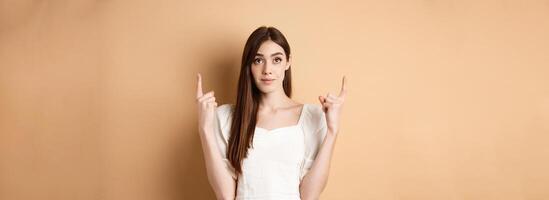Hopeful romantic girl in white dress pointing fingers up, showing empty space for company logo, standing on beige background photo