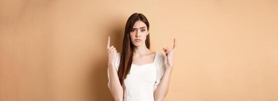 Skeptical woman frowning and pointing fingers up, having doubts about product, standing on beige background photo