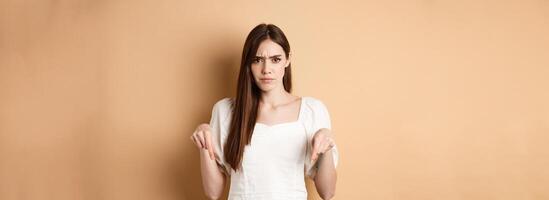 Skeptical young woman frowning, pointing hands down at something bad or disappointing, look upset, standing on beige background photo