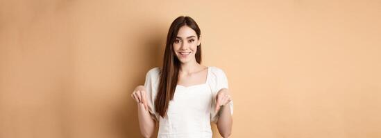 Cheerful young girl with lovely smile, pointing fingers down, making announcement or inviting click link, showing logo, beige background photo