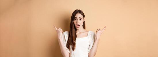 Surprised young girl pointing sideways at left and right, gasping and saying wow amazed, showing two cool choices, beige background photo