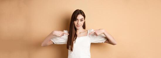 Young serious woman pointing at center, showing logo with confident face, standing in dress on beige background photo