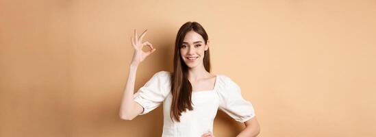Very good. Smiling cute woman in white blouse showing okay sign in approval, like and praise excellent choice, standing satisfied on beige background photo