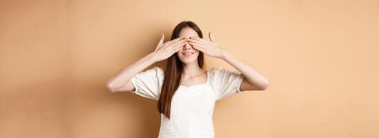 Happy romantic girl covering eyes and waiting for surprise, smiling while anticipating gift, standing on beige background photo