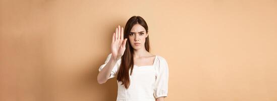 Nope stop right there. Serious and confident woman stretch out hand to forbid something, frowning and saying no, disagree and reject bad offer, standing on beige background photo