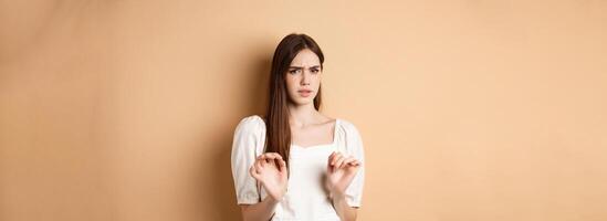 Stay away from me. Disgusted young woman frowning and looking with aversion at something stinky, rejecting disgusting thing, standing on beige background photo