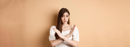 Girl says no. Skeptical young woman disagree or prohibit action, showing cross stop gesture, make nope sign, disapprove bad idea, standing on beige background photo