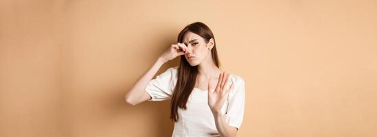 Displeased woman plug her nose from bad smell, showing stop gesture and frowning from disgust. Shut nostrils in aversion, standing on beige background photo