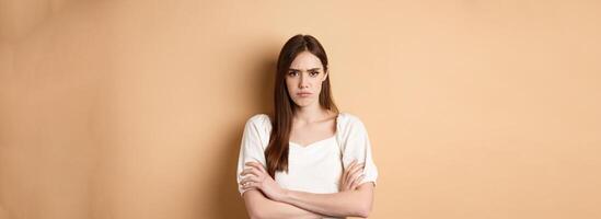 Angry girlfriend cross arms on chest and frowning, looking offended or disappointed, sulking at you, standing mad on beige background photo