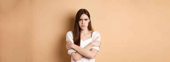 Offended woman hugging herself and frowning upset, feeling timid and defensive, standing on beige background photo