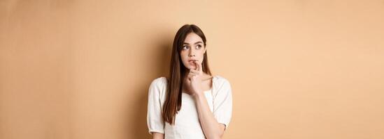 Nervous young girl biting fingernail and looking aside excited, making choice, thinking about something, standing on beige background photo