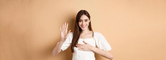Cute smiling girl make promise, put hand on heart and tell truth, being honest, swearing to you, standing on beige background photo