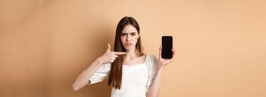 Disappointed frowning girl pointing at empty phone screen, complaining at online news, standing on beige background photo