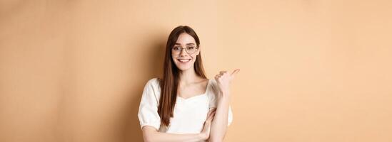Cheerful woman in eyewear pointing finger left at logo, smiling happy, standing on beige background photo