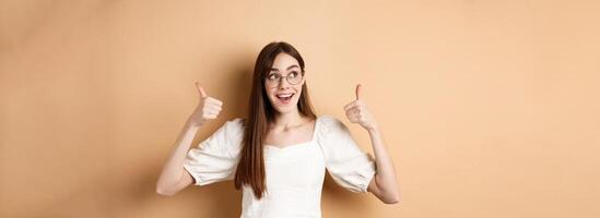 Happy girl in glasses show thumbs up, looking aside at good logo, standing satisfied on beige background photo
