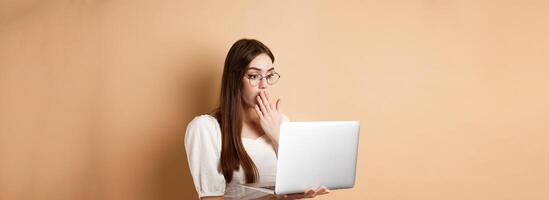 Shocked girl look at laptop screen, covering mouth with amazed face, standing in glasses on beige background photo