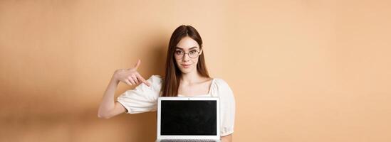 Confident young woman in glasses pointing at laptop screen, showing computer display, standing on beige background photo