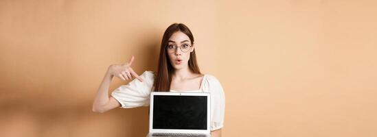 Excited girl in glasses making presentation on computer, pointing hand at laptop screen and say wow, standing on beige background photo