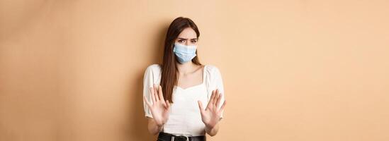 Pandemic and healthcare concept. Keep distance. Young woman in medical mask stretch out hand in stop sign, telling to stay away and frowning disgusted, beige background photo