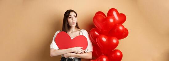 Valentines day and love concept. Intrigued tender girl hugging big red heart cutout and stand near balloons, looking aside at logo, saying wow, beige background photo