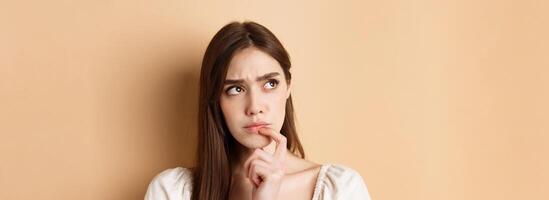 Portrait of thoughtful girl frowning and looking aside pensive, thinking or making choice, standing on beige background photo