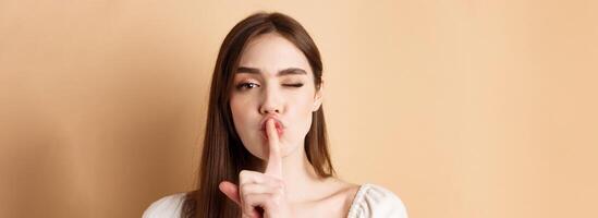 Close up of attractive woman shushing at camera, hiding secret, making hush sign and winking, show taboo sign, standing on beige background photo