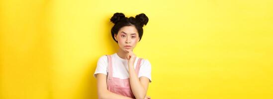 Suspicious asian girl looking at camera and thinking, stare with disbelief, standing with makeup on yellow background photo