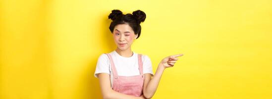 Glamour asian girl with bright makeup, pointing finger left and winking at camera, hint on good promo deal, standing on yellow background photo