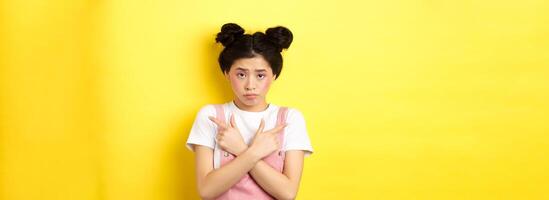 Indecisive timid asian girl pointing sideways, showing left and right ways and sulking upset, standing sad on yellow background photo