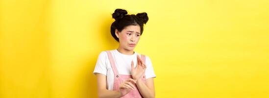 Disgusted asian girl raising hands up defensive, block something disgusting, turn away from aversion and reluctance, standing on yellow background photo