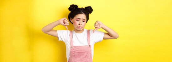 Asian beauty girl cringe from something disgusting, look away and showing thumbs-down to express dislike, negative emotions, standing in summer clothes on yellow background photo
