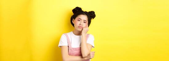 Sad and bored teen asian girl standing alone, looking at camera indifferent, staring with boredom, yellow background photo