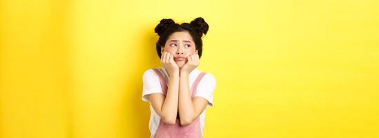 Sad moody asian girl, lean face on hands, looking left with sulking expression, standing in summer clothes on yellow background photo